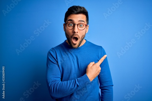 Young handsome man with beard wearing casual sweater and glasses over blue background Surprised pointing with finger to the side, open mouth amazed expression. photo