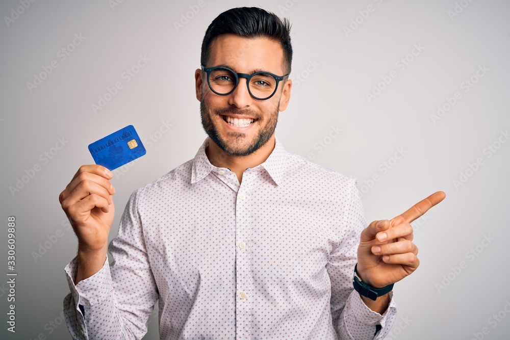 Young business man holding credit card over isolated background very happy pointing with hand and finger to the side Young business man holding credit card over isolated background very happy pointing with hand and finger to the side