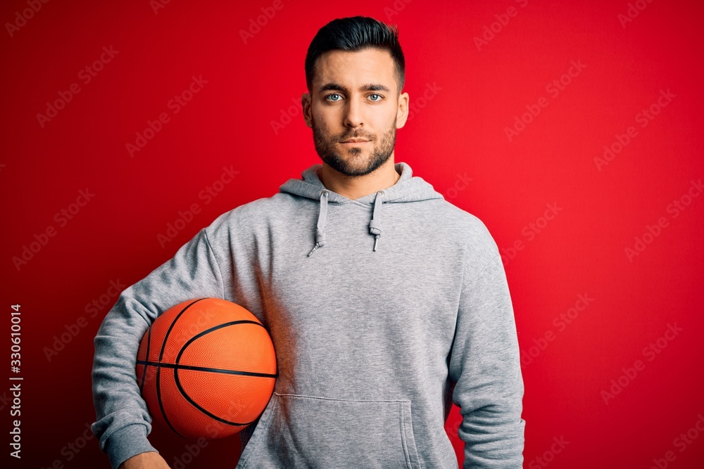 Young sports man holding basketball ball over red isolated background with a confident expression on smart face thinking serious