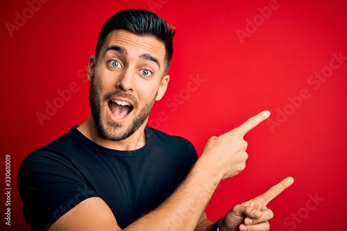Young handsome man wearing casual black t-shirt standing over isolated red background smiling and looking at the camera pointing with two hands and fingers to the side.