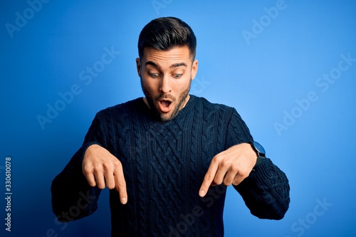 Young handsome man wearing casual sweater standing over isolated blue background Pointing down with fingers showing advertisement, surprised face and open mouth