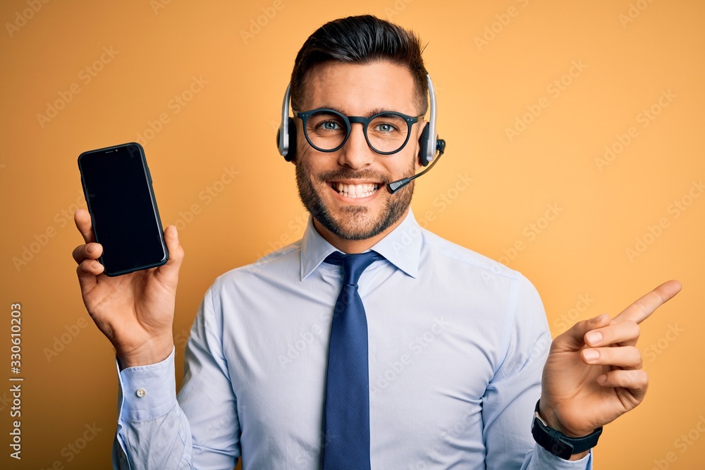 Business operator man with customer service headset from call center showing smartphone screen very happy pointing with hand and finger to the side Business operator man with customer service headset from call center showing smartphone screen very happy pointing with hand and finger to the side