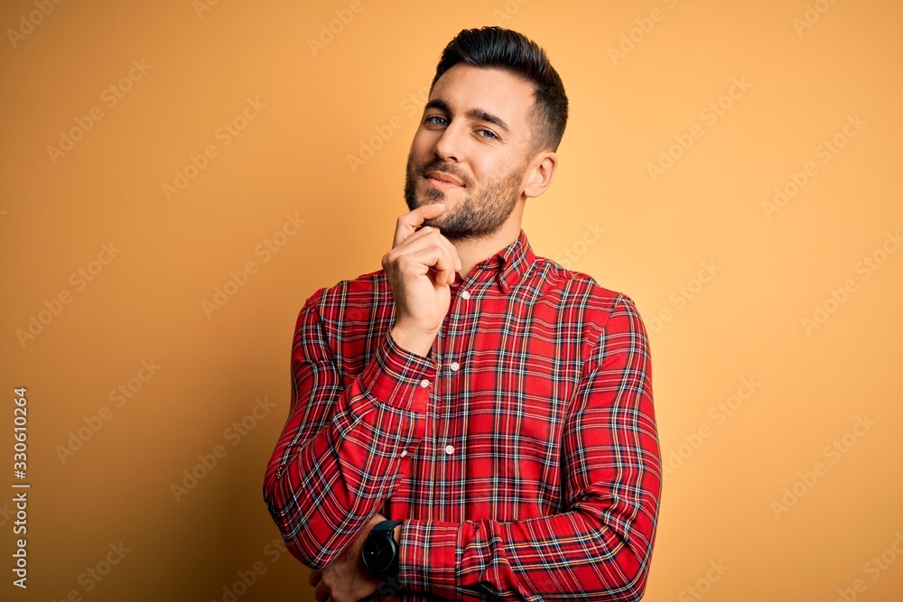 Young handsome man wearing casual shirt standing over isolated yellow background looking confident at the camera with smile with crossed arms and hand raised on chin. Thinking positive. Young handsome man wearing casual shirt standing over isolated yellow background looking confident at the camera with smile with crossed arms and hand raised on chin. Thinking positive.