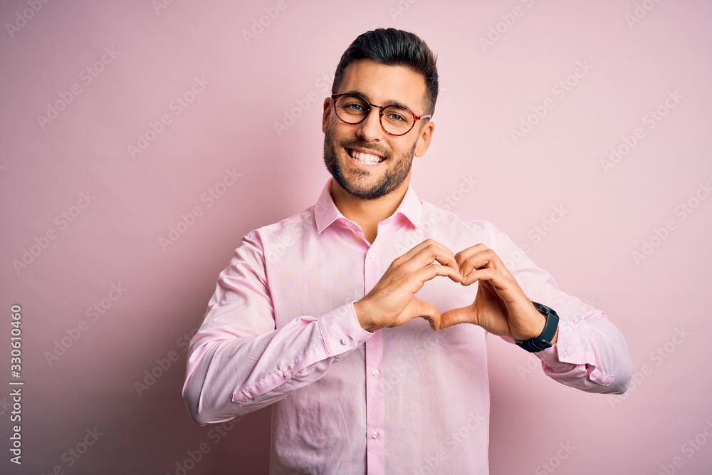 Young handsome man wearing elegant shirt and glasses standing over pink background smiling in love showing heart symbol and shape with hands. Romantic concept. Young handsome man wearing elegant shirt and glasses standing over pink background smiling in love showing heart symbol and shape with hands. Romantic concept.