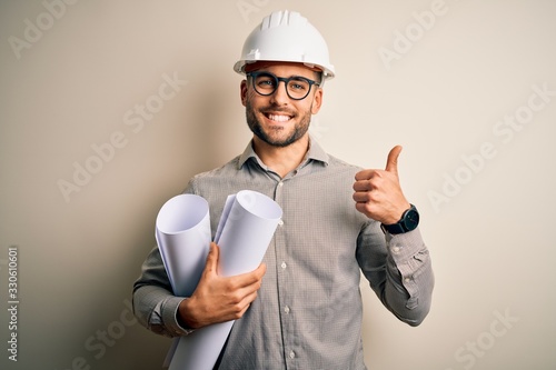 Young architect man wearing contractor helmet holding project paper plan over isolated background happy with big smile doing ok sign, thumb up with fingers, excellent sign photo