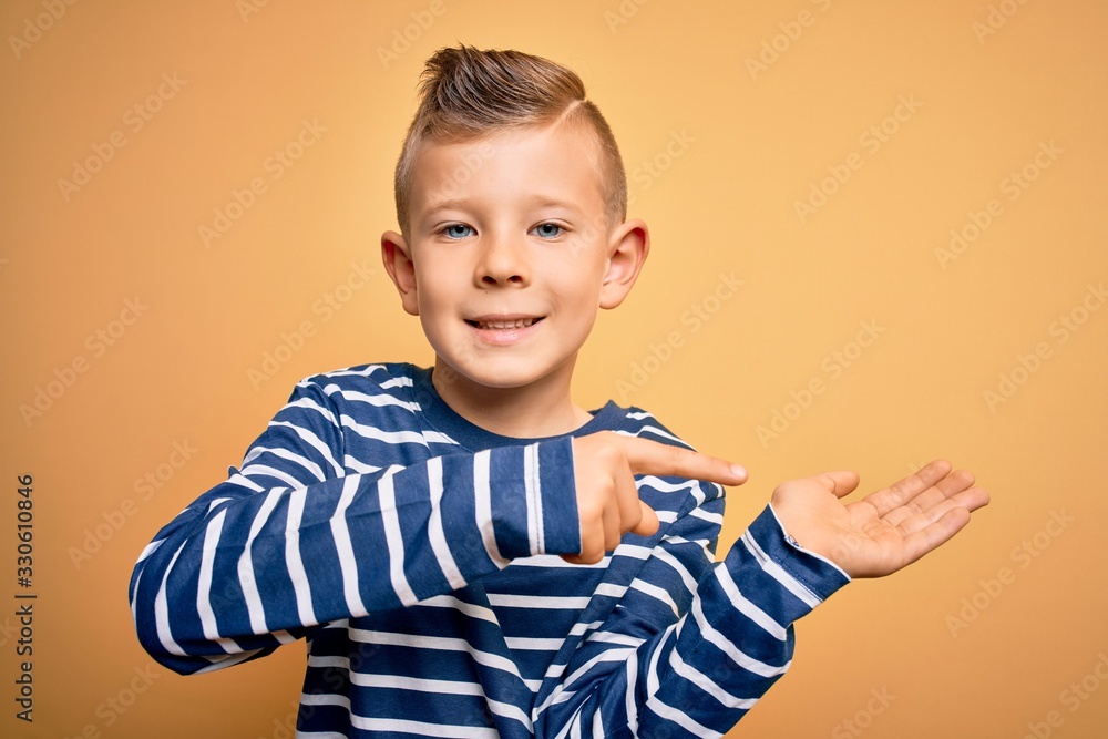 Young little caucasian kid with blue eyes wearing nautical striped shirt over yellow background amazed and smiling to the camera while presenting with hand and pointing with finger. Young little caucasian kid with blue eyes wearing nautical striped shirt over yellow background amazed and smiling to the camera while presenting with hand and pointing with finger.