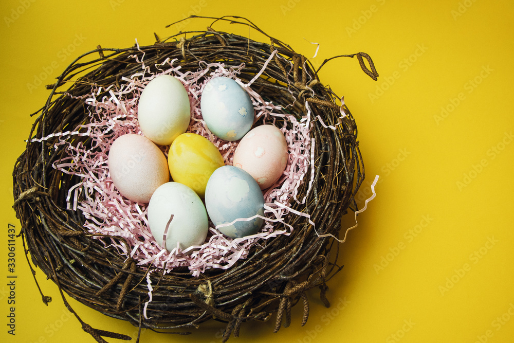 A nest of twigs and hay and a pink paper filler with pastel colored Easter eggs. Colored eggs for the holiday of light Easter. Easter card on a yellow background A nest of twigs and hay and a pink paper filler with pastel colored Easter eggs. Colored eggs for the holiday of light Easter. Easter card on a yellow background