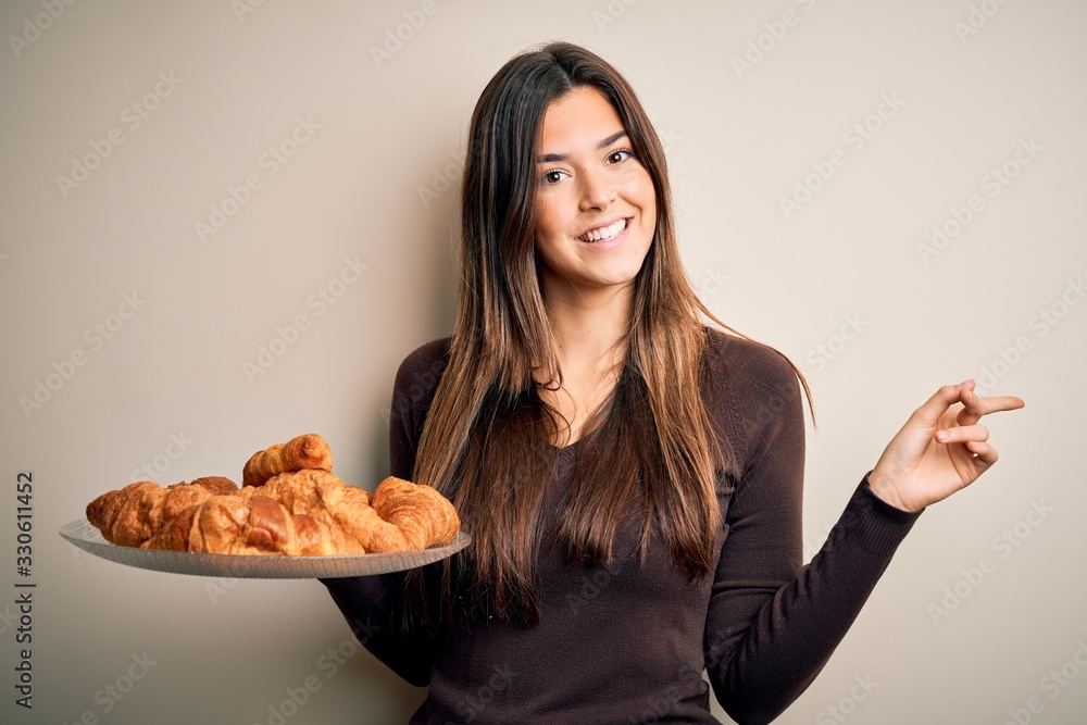 Young beautiful girl holding plate with sweet croissants for breakfast over white background very happy pointing with hand and finger to the side Young beautiful girl holding plate with sweet croissants for breakfast over white background very happy pointing with hand and finger to the side