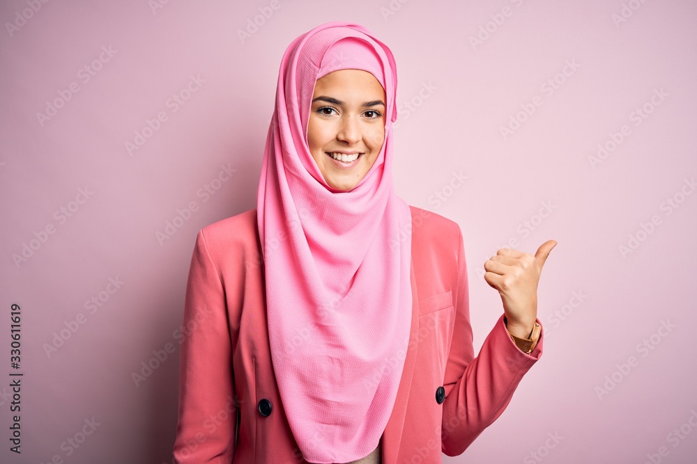 Young beautiful girl wearing muslim hijab standing over isolated pink background smiling with happy face looking and pointing to the side with thumb up. Young beautiful girl wearing muslim hijab standing over isolated pink background smiling with happy face looking and pointing to the side with thumb up.