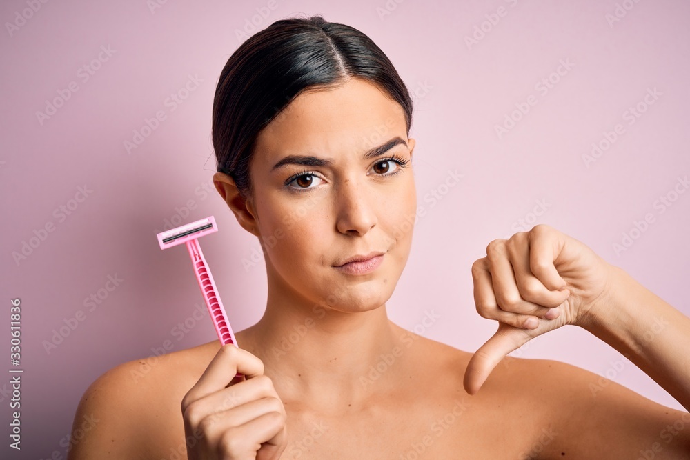 Young beautiful girl using shaver for depilation standing over isolated pink background with angry face, negative sign showing dislike with thumbs down, rejection concept