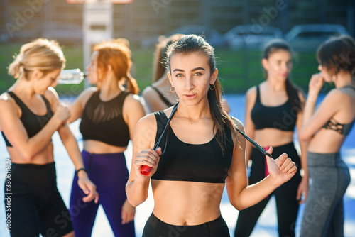 Pretty young woman with jump rope on her neck looking at camera after outdoors workout with her female friends on the background on the stadium. © gorynvd