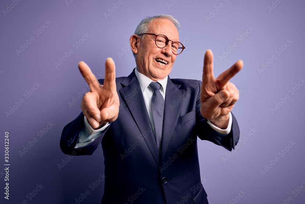 Grey haired senior business man wearing glasses and elegant suit and tie over purple background smiling with tongue out showing fingers of both hands doing victory sign. Number two. Grey haired senior business man wearing glasses and elegant suit and tie over purple background smiling with tongue out showing fingers of both hands doing victory sign. Number two.