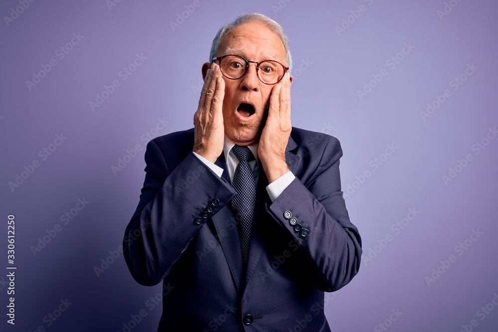 Grey haired senior business man wearing glasses and elegant suit and tie over purple background afraid and shocked, surprise and amazed expression with hands on face Grey haired senior business man wearing glasses and elegant suit and tie over purple background afraid and shocked, surprise and amazed expression with hands on face