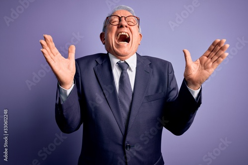 Grey haired senior business man wearing glasses and elegant suit and tie over purple background celebrating mad and crazy for success with arms raised and closed eyes screaming excited. Winner concept