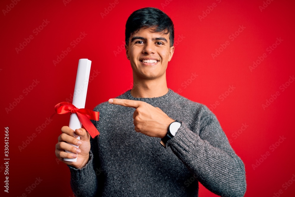 Young student man holding university graduated diploma degree over red background very happy pointing with hand and finger Young student man holding university graduated diploma degree over red background very happy pointing with hand and finger