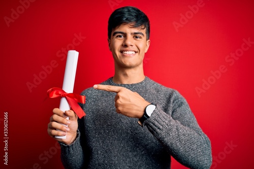 Young student man holding university graduated diploma degree over red background very happy pointing with hand and finger photo