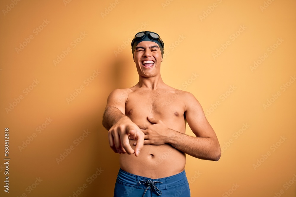 Young handsome man shirtless wearing swimsuit and swim cap over isolated yellow background laughing at you, pointing finger to the camera with hand over body, shame expression