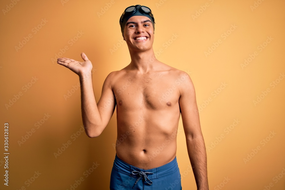 Young handsome man shirtless wearing swimsuit and swim cap over isolated yellow background smiling cheerful presenting and pointing with palm of hand looking at the camera.