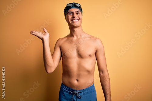 Young handsome man shirtless wearing swimsuit and swim cap over isolated yellow background smiling cheerful presenting and pointing with palm of hand looking at the camera. © Krakenimages.com