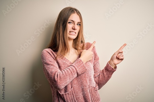 Young beautiful redhead woman wearing pink casual sweater over isolated white background Pointing aside worried and nervous with both hands, concerned and surprised expression