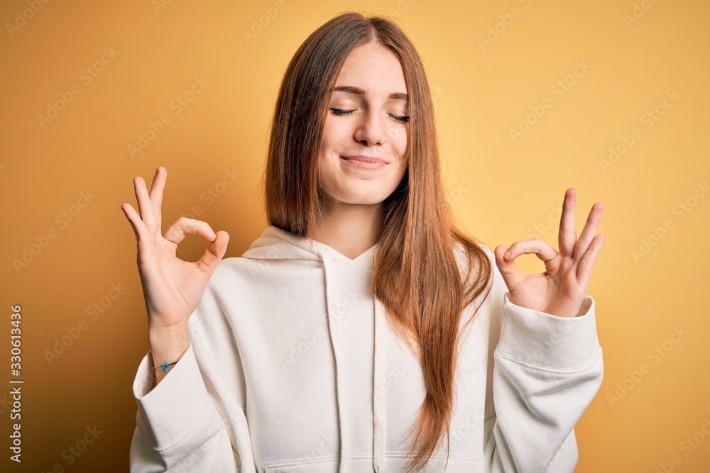 Young beautiful redhead sporty woman wearing sweatshirt over isolated yellow background relax and smiling with eyes closed doing meditation gesture with fingers. Yoga concept. Young beautiful redhead sporty woman wearing sweatshirt over isolated yellow background relax and smiling with eyes closed doing meditation gesture with fingers. Yoga concept.