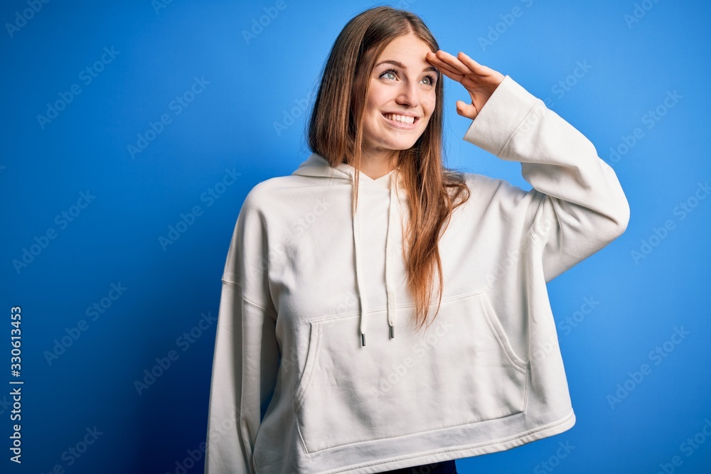 Young beautiful redhead sporty woman wearing sweatshirt over isolated blue background very happy and smiling looking far away with hand over head. Searching concept. Young beautiful redhead sporty woman wearing sweatshirt over isolated blue background very happy and smiling looking far away with hand over head. Searching concept.