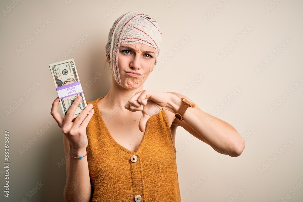 Young woman injured wearing bandage on head holding money from medical insurance payment with angry face, negative sign showing dislike with thumbs down, rejection concept Young woman injured wearing bandage on head holding money from medical insurance payment with angry face, negative sign showing dislike with thumbs down, rejection concept