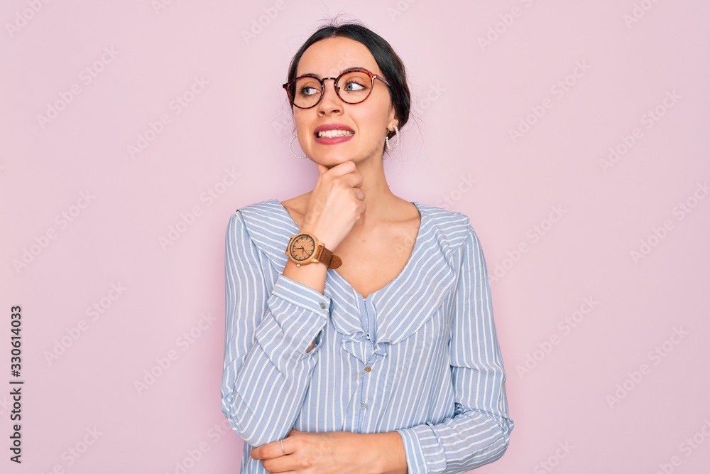 Young beautiful woman wearing casual striped shirt and glasses over pink background with hand on chin thinking about question, pensive expression. Smiling with thoughtful face. Doubt concept.
