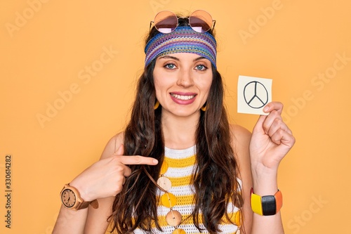 Beautiful hippie woman with blue eyes wearing sunnglasses holding peace symbol reminder with surprise face pointing finger to himself photo