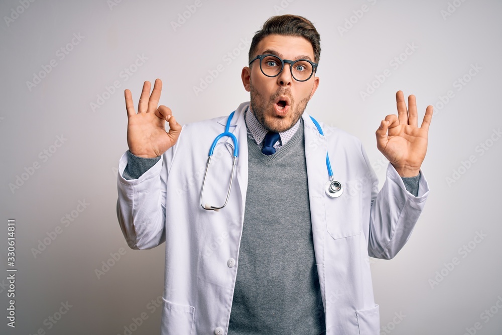 Young doctor man with blue eyes wearing medical coat and stethoscope over isolated background looking surprised and shocked doing ok approval symbol with fingers. Crazy expression Young doctor man with blue eyes wearing medical coat and stethoscope over isolated background looking surprised and shocked doing ok approval symbol with fingers. Crazy expression