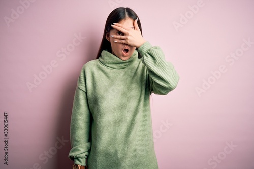 Young beautiful asian woman wearing green winter sweater over pink solated background peeking in shock covering face and eyes with hand, looking through fingers with embarrassed expression. © Krakenimages.com