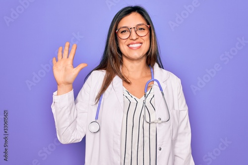 Professional doctor woman wearing stethoscope and medical coat over purple background showing and pointing up with fingers number five while smiling confident and happy.
