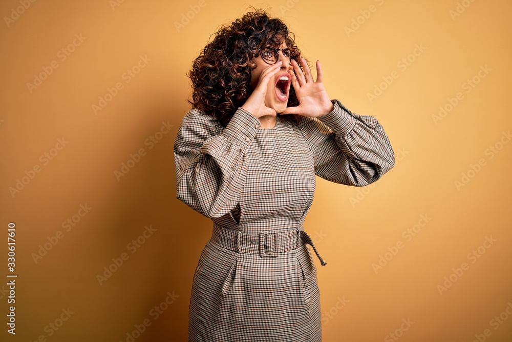 Beautiful arab business woman wearing dress and glasses standing over yellow background Shouting angry out loud with hands over mouth