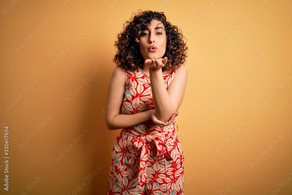 Young beautiful curly arab woman on vacation wearing summer floral dress and sunglasses looking at the camera blowing a kiss with hand on air being lovely and sexy. Love expression. Young beautiful curly arab woman on vacation wearing summer floral dress and sunglasses looking at the camera blowing a kiss with hand on air being lovely and sexy. Love expression.