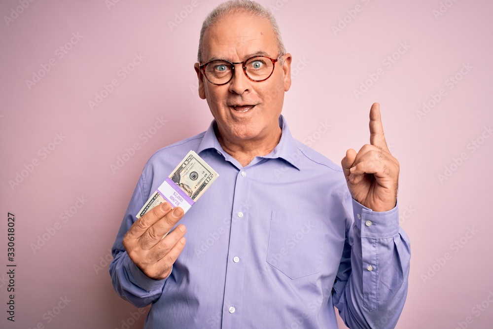 Middle age hoary man holding bunch of dollars banknotes over isolated pink background surprised with an idea or question pointing finger with happy face, number one Middle age hoary man holding bunch of dollars banknotes over isolated pink background surprised with an idea or question pointing finger with happy face, number one