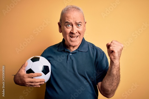 Middle age hoary player man playing soccer holding football ball over yellow background screaming proud and celebrating victory and success very excited, cheering emotion © Krakenimages.com
