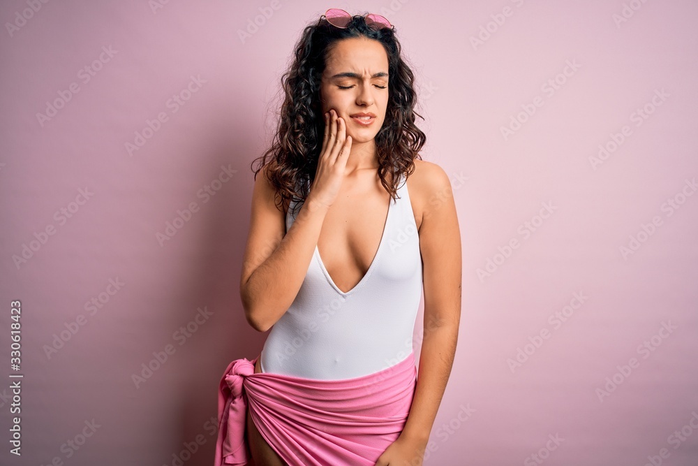 Beautiful woman with curly hair on vacation wearing white swimsuit over pink background touching mouth with hand with painful expression because of toothache or dental illness on teeth. Dentist Beautiful woman with curly hair on vacation wearing white swimsuit over pink background touching mouth with hand with painful expression because of toothache or dental illness on teeth. Dentist