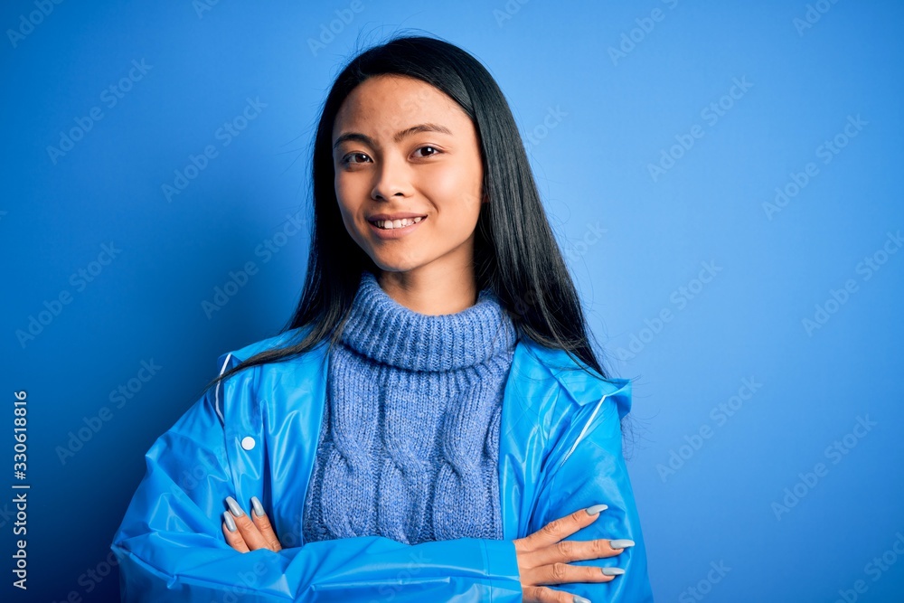 Young beautiful chinese woman wearing rain coat standing over isolated blue background happy face smiling with crossed arms looking at the camera. Positive person. Young beautiful chinese woman wearing rain coat standing over isolated blue background happy face smiling with crossed arms looking at the camera. Positive person.