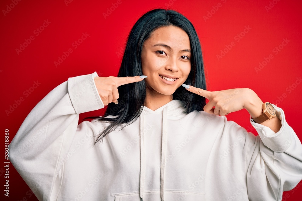Young beautiful chinese sporty woman wearing sweatshirt over isolated red background smiling cheerful showing and pointing with fingers teeth and mouth. Dental health concept. Young beautiful chinese sporty woman wearing sweatshirt over isolated red background smiling cheerful showing and pointing with fingers teeth and mouth. Dental health concept.