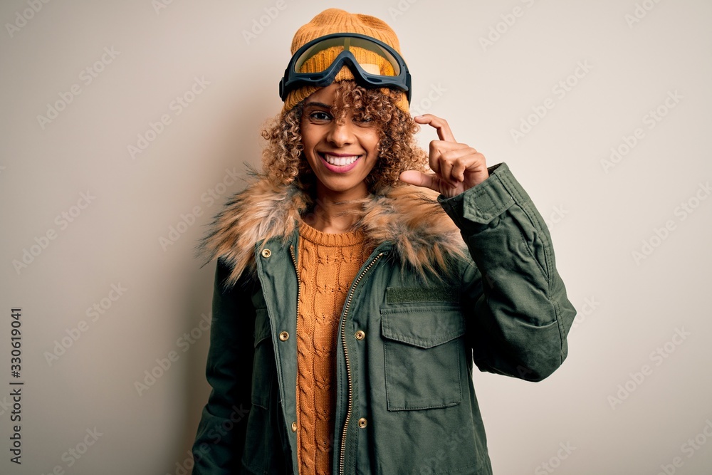 Young african american skier woman with curly hair wearing snow sportswear and ski goggles smiling and confident gesturing with hand doing small size sign with fingers looking and the camera. Measure Young african american skier woman with curly hair wearing snow sportswear and ski goggles smiling and confident gesturing with hand doing small size sign with fingers looking and the camera. Measure