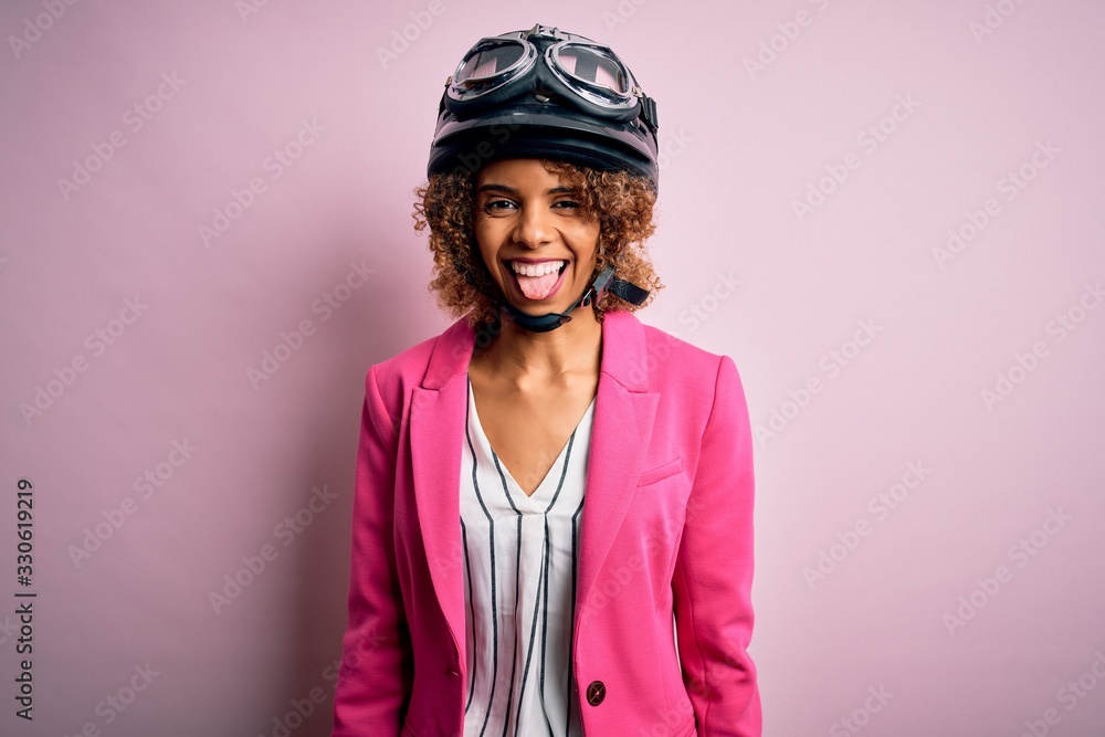 African american motorcyclist woman with curly hair wearing moto helmet over pink background sticking tongue out happy with funny expression. Emotion concept.