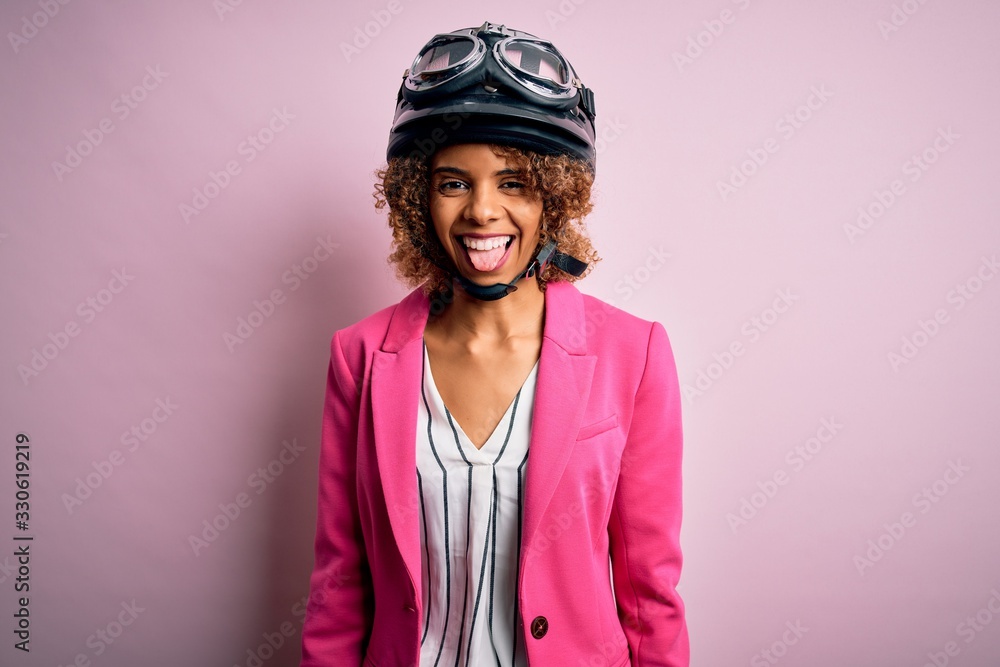 African american motorcyclist woman with curly hair wearing moto helmet over pink background sticking tongue out happy with funny expression. Emotion concept. African american motorcyclist woman with curly hair wearing moto helmet over pink background sticking tongue out happy with funny expression. Emotion concept.