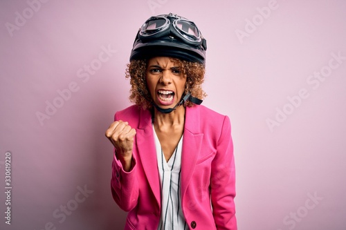 African american motorcyclist woman with curly hair wearing moto helmet over pink background angry and mad raising fist frustrated and furious while shouting with anger. Rage and aggressive concept. © Krakenimages.com