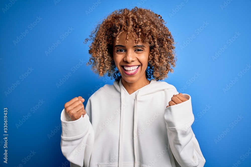 Beautiful african american sporty woman wearing casual sweatshirt over blue background celebrating surprised and amazed for success with arms raised and open eyes. Winner concept. Beautiful african american sporty woman wearing casual sweatshirt over blue background celebrating surprised and amazed for success with arms raised and open eyes. Winner concept.