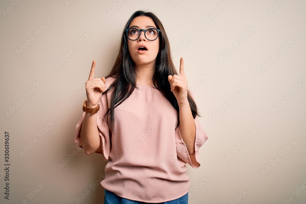 Young brunette elegant woman wearing glasses over isolated background amazed and surprised looking up and pointing with fingers and raised arms. Young brunette elegant woman wearing glasses over isolated background amazed and surprised looking up and pointing with fingers and raised arms.