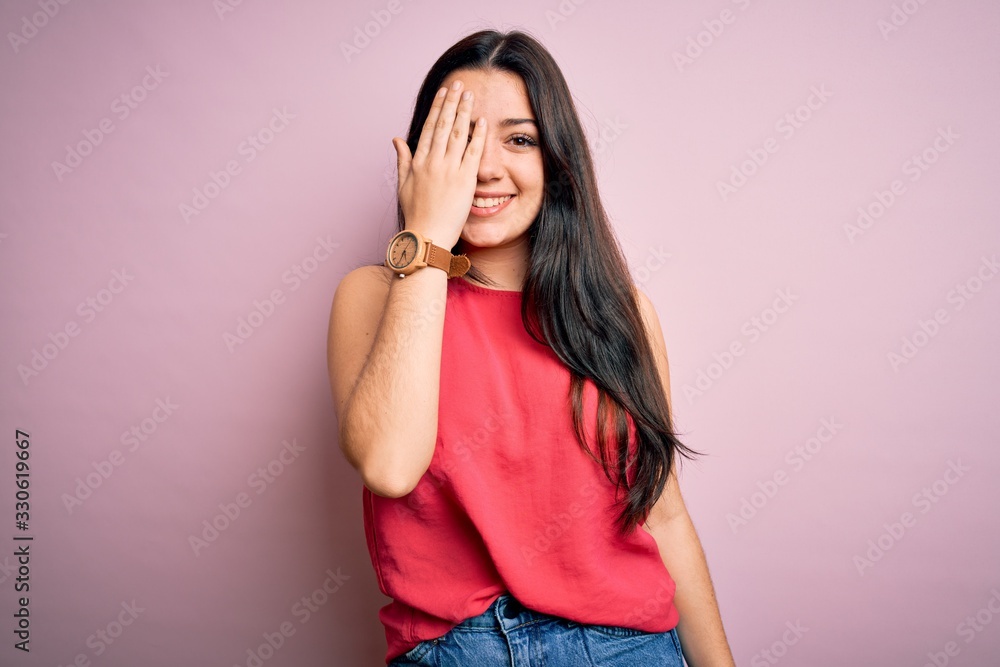 Young brunette woman wearing casual summer shirt over pink isolated background covering one eye with hand, confident smile on face and surprise emotion. Young brunette woman wearing casual summer shirt over pink isolated background covering one eye with hand, confident smile on face and surprise emotion.