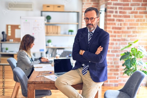 Middle age handsome businessman wearing glasses sitting on desk at the office skeptic and nervous, disapproving expression on face with crossed arms. Negative person.
