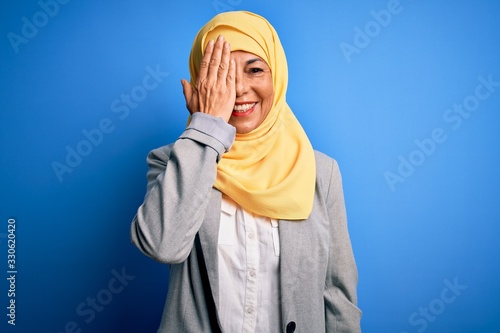 Middle age brunette business woman wearing muslim traditional hijab over blue background covering one eye with hand, confident smile on face and surprise emotion.