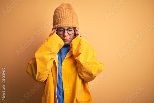 Middle age woman wearing yellow raincoat and winter hat over isolated background with hand on head for pain in head because stress. Suffering migraine. © Krakenimages.com