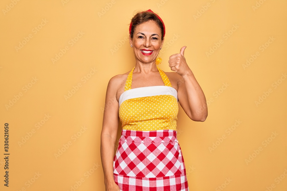 Middle age senior pin up housewife woman wearing 50s style retro dress and apron doing happy thumbs up gesture with hand. Approving expression looking at the camera showing success. Middle age senior pin up housewife woman wearing 50s style retro dress and apron doing happy thumbs up gesture with hand. Approving expression looking at the camera showing success.